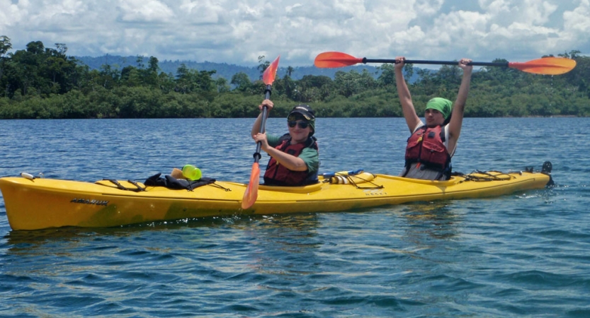 Two people wearing life jackets sit tandem in a yellow kayak. One of them is holding the paddle in the air in celebration. 